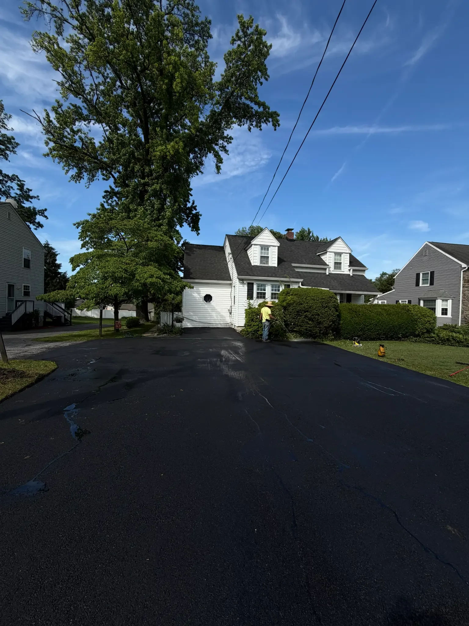 Freshly sealcoated residential driveway in front of a white two-story home, with a crew member finishing the edge by hand